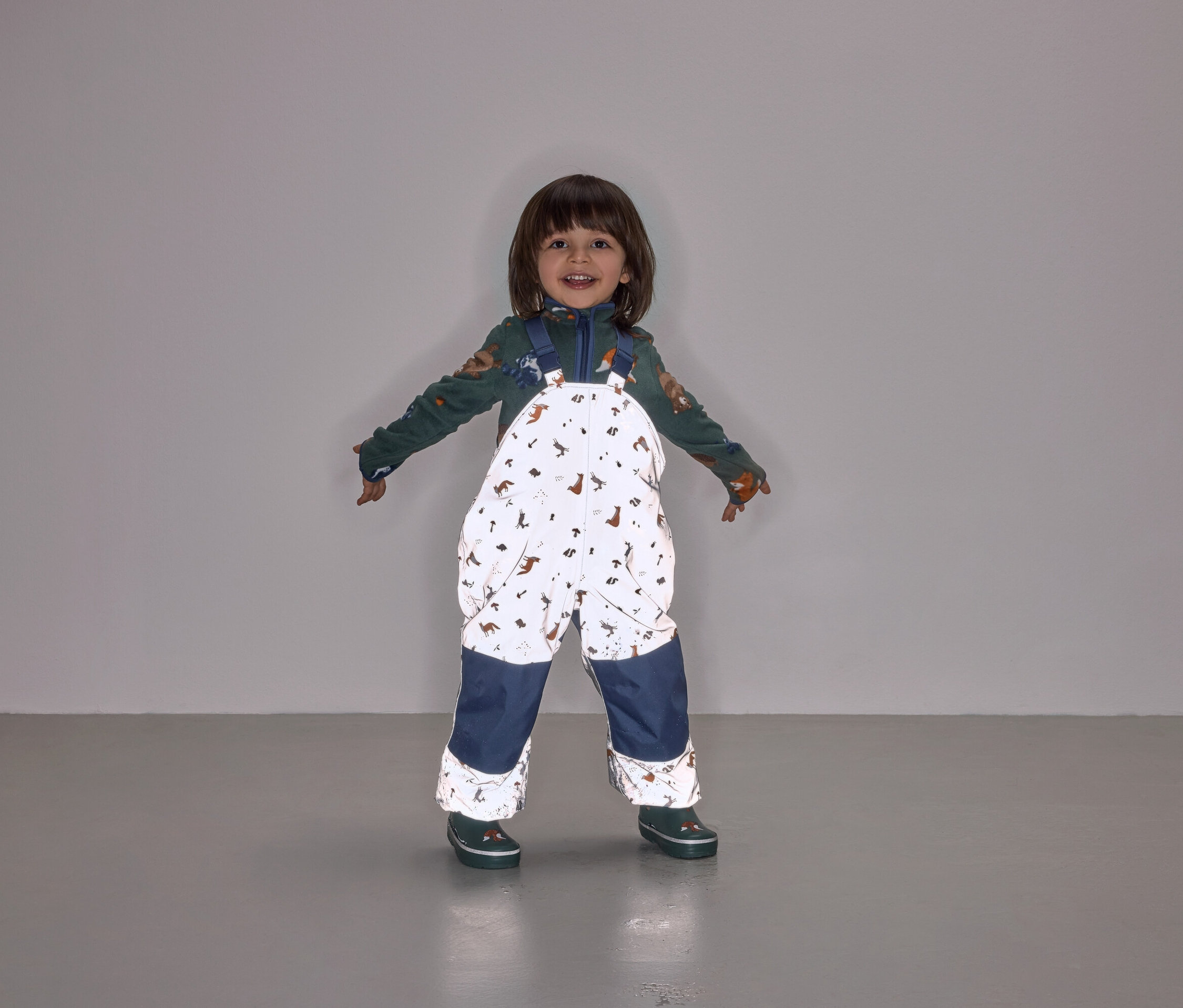 Usmívající se dítě stojí s rozpaženýma rukama a má na sobě kombinézu s laclem s potiskem zvířat a tmavě modrými nohavicemi.