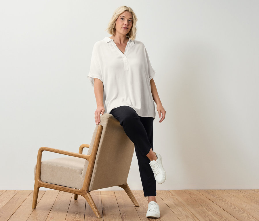 Blonďatá žena opřená o béžové křeslo pózuje v krémově bílé halence.