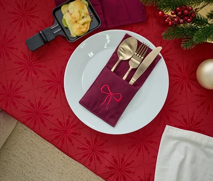 Talíř s ubrouskem s výšivkou, zlatý příbor, raclette pánev a vánoční dekorace na červeném ubrusu.