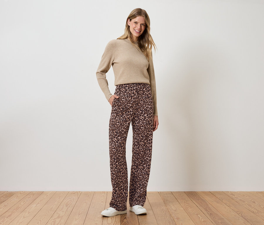 Žena stojí a má ruce v kapsách hnědých květovaných kalhot a béžový svetr.
