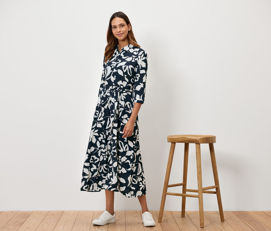Žena stojí vedle dřevěné stoličky a má na sobě tmavě modré šaty s bílým květinovým vzorem.