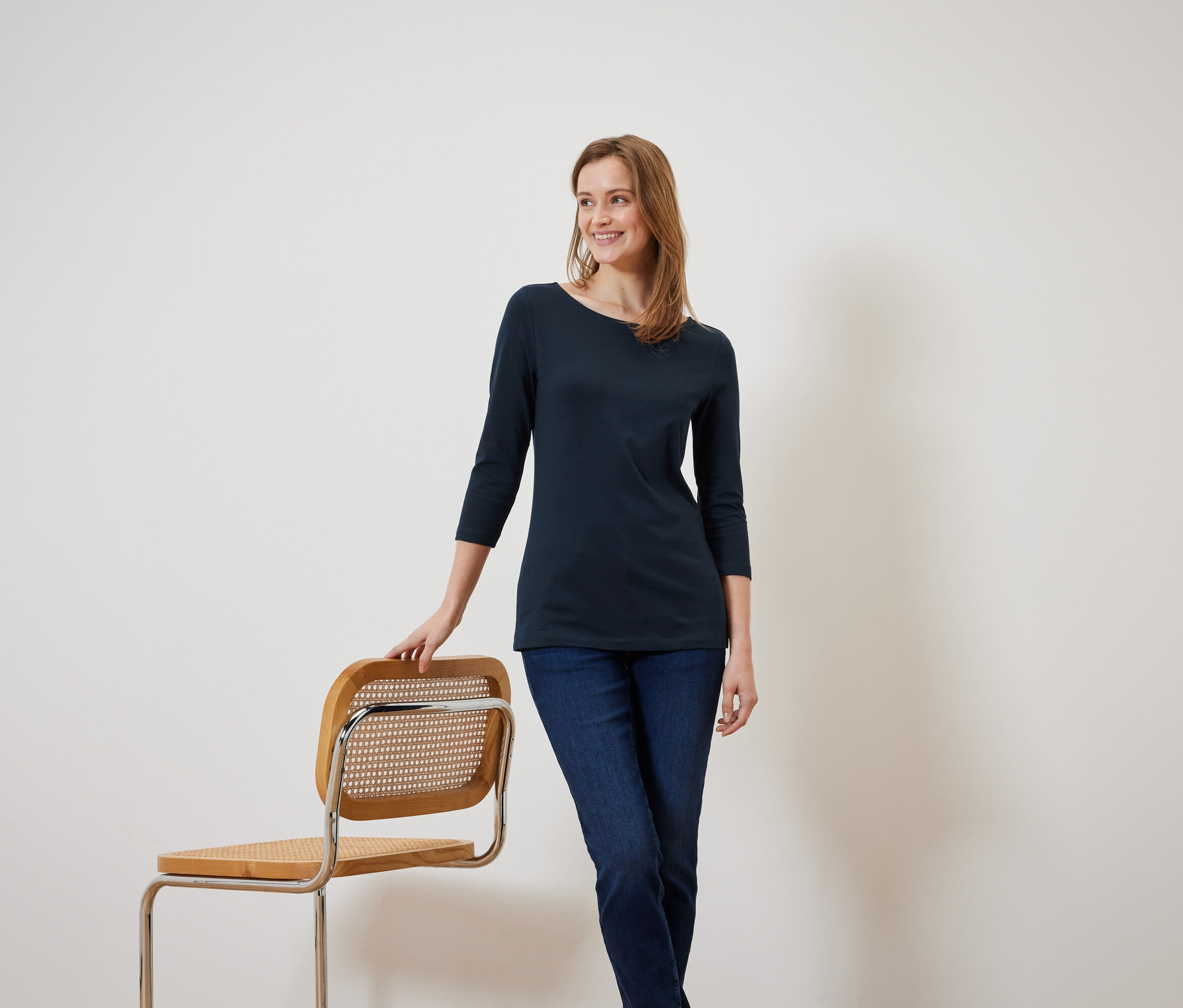 Usmívající se žena v modré blůze s tříčtvrtečním rukávem se opírá o dřevěnou židli.