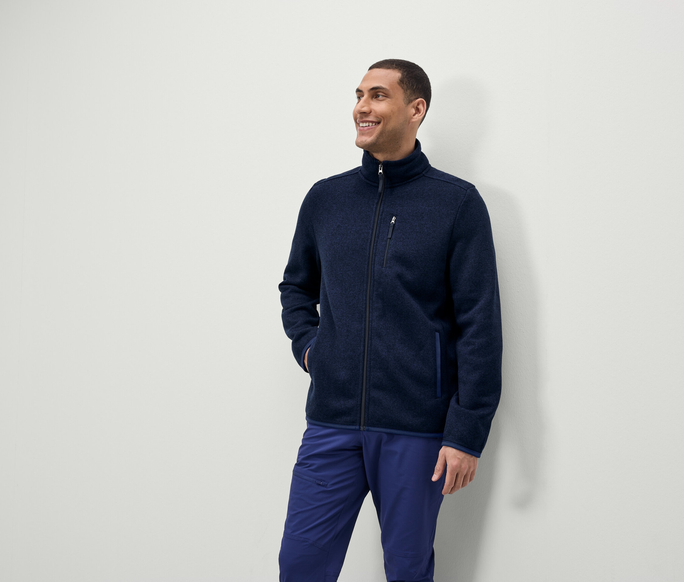 Muž se usmívá a dívá se stranou, má na sobě modrou fleecovou bundu na zip a modré kalhoty.