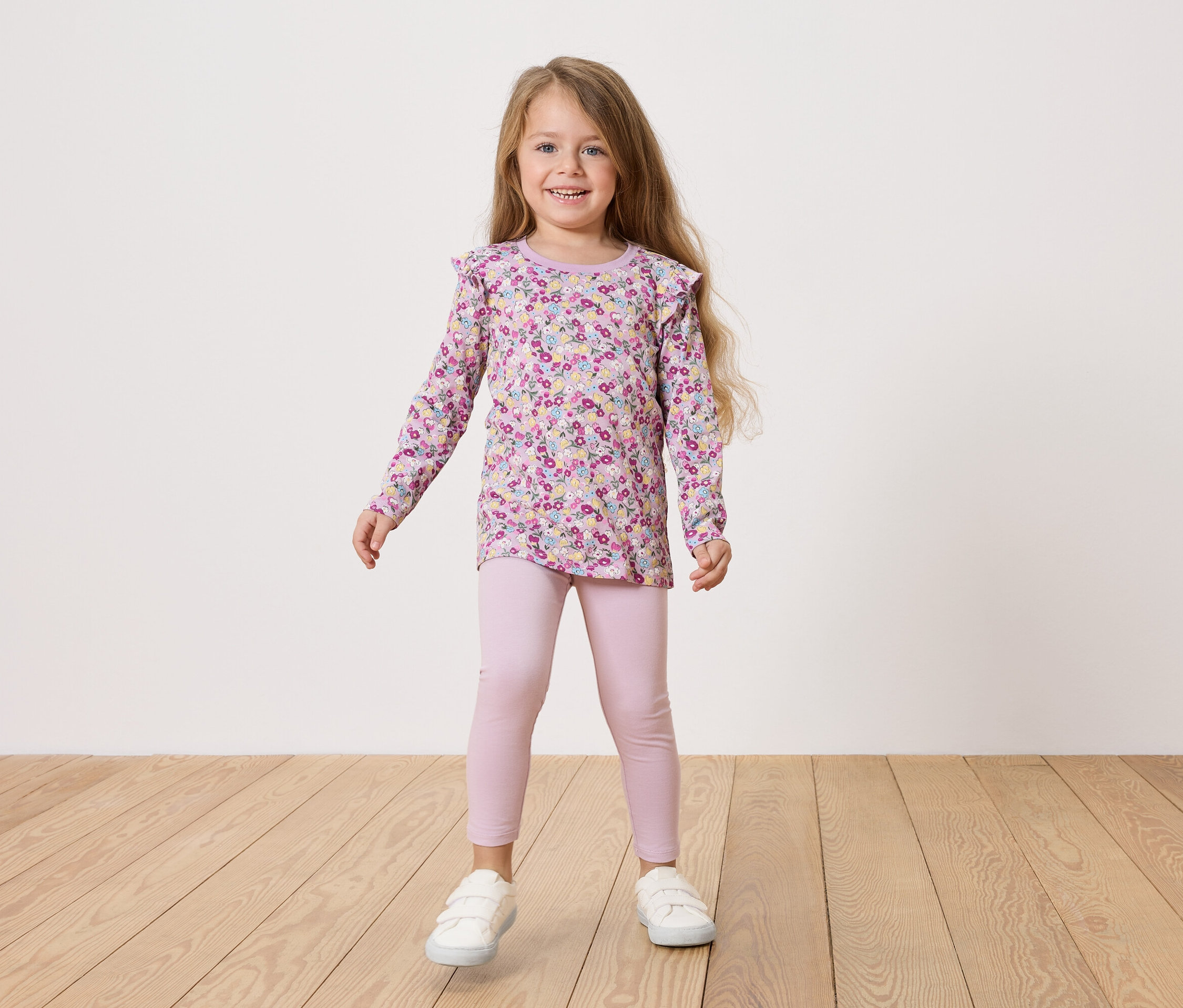 Dívka s blond vlasy má na sobě květinovou blůzu, růžové legíny a bílé boty.