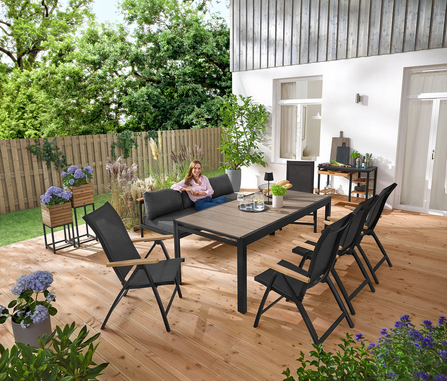 Žena sedí na zahradní lavici 2 v 1 – lehátku »Elin«. V pozadí stojí Stojany na květináče »Elin«, 2 ks. Na terase je také grilovací vozík s polycementovou pracovní plochou a rozkládací zahradní stůl se stolní deskou Duraboard.