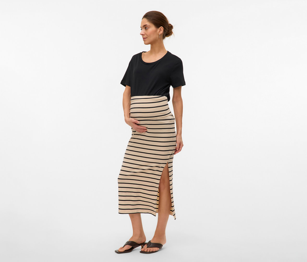 Žena v černém tričku a pruhované maxi sukni MAMALICIOUS »MLEVA« s rozparkem. Drží ruce na břiše.