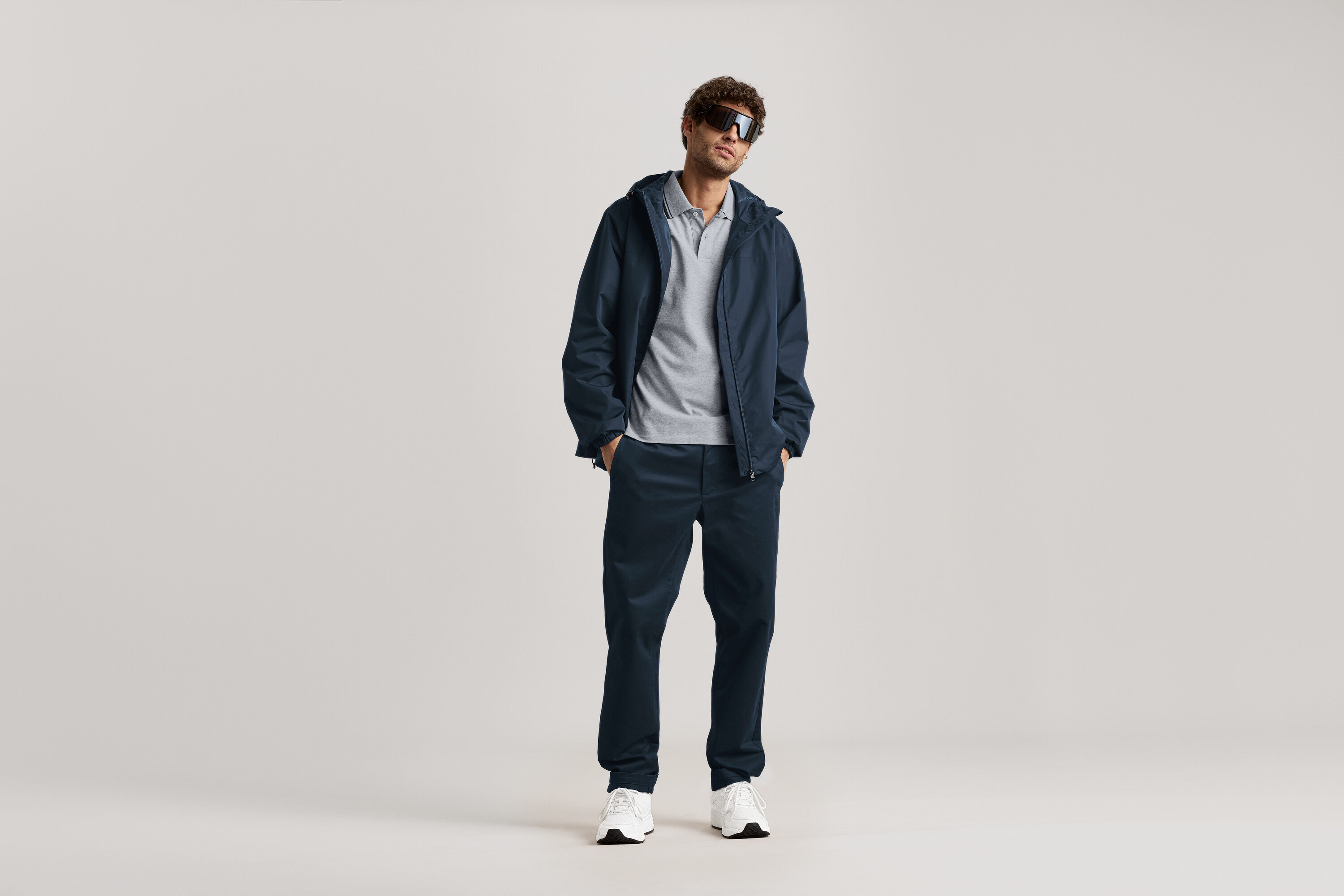 Muž stojí se slunečními brýlemi, tmavě modrou bundou a tmavě modrými chino kalhotami.