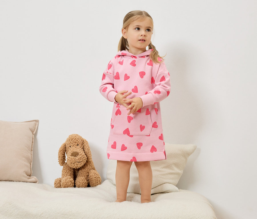 Malá dívka s blond copánky stojí na posteli a má na sobě růžové šaty s kapucí a červenými srdíčky. Vedle ní sedí plyšový pes.