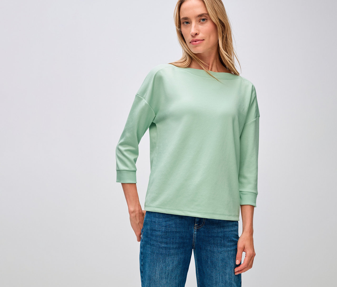 Žena má na sobě tričko Street One s žebrovaným detailem, šalvějově zelené a džíny.