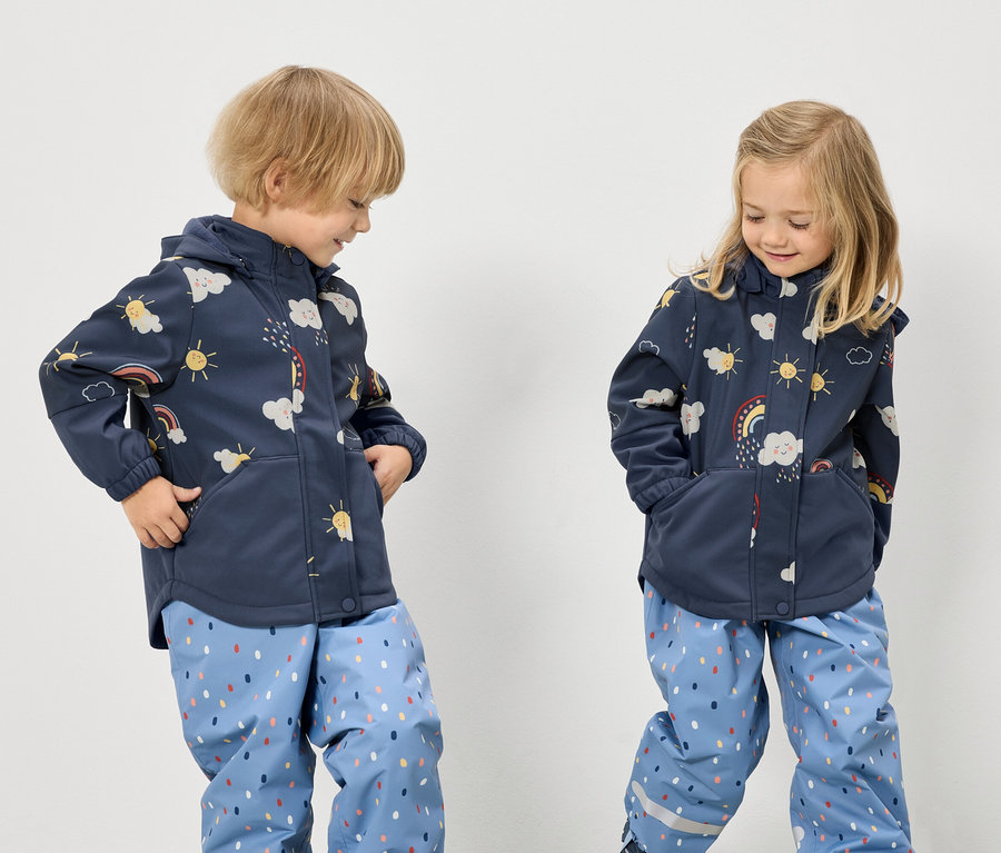 Dvě děti v modrých bundách se vzorem duhy, mraků a slunce a modrých kalhotách s puntíkovaným vzorem stojí na bílém pozadí.