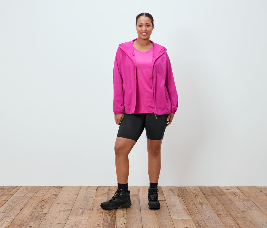 Žena stojí a má na sobě růžovou bundu, růžové tričko, krátké černé kraťasy, černé ponožky a černé boty.