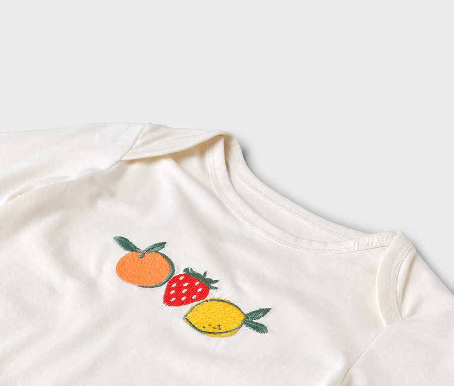 Detailní záběr na bílé dětské body s výšivkou mandarinky, jahody a citronu.