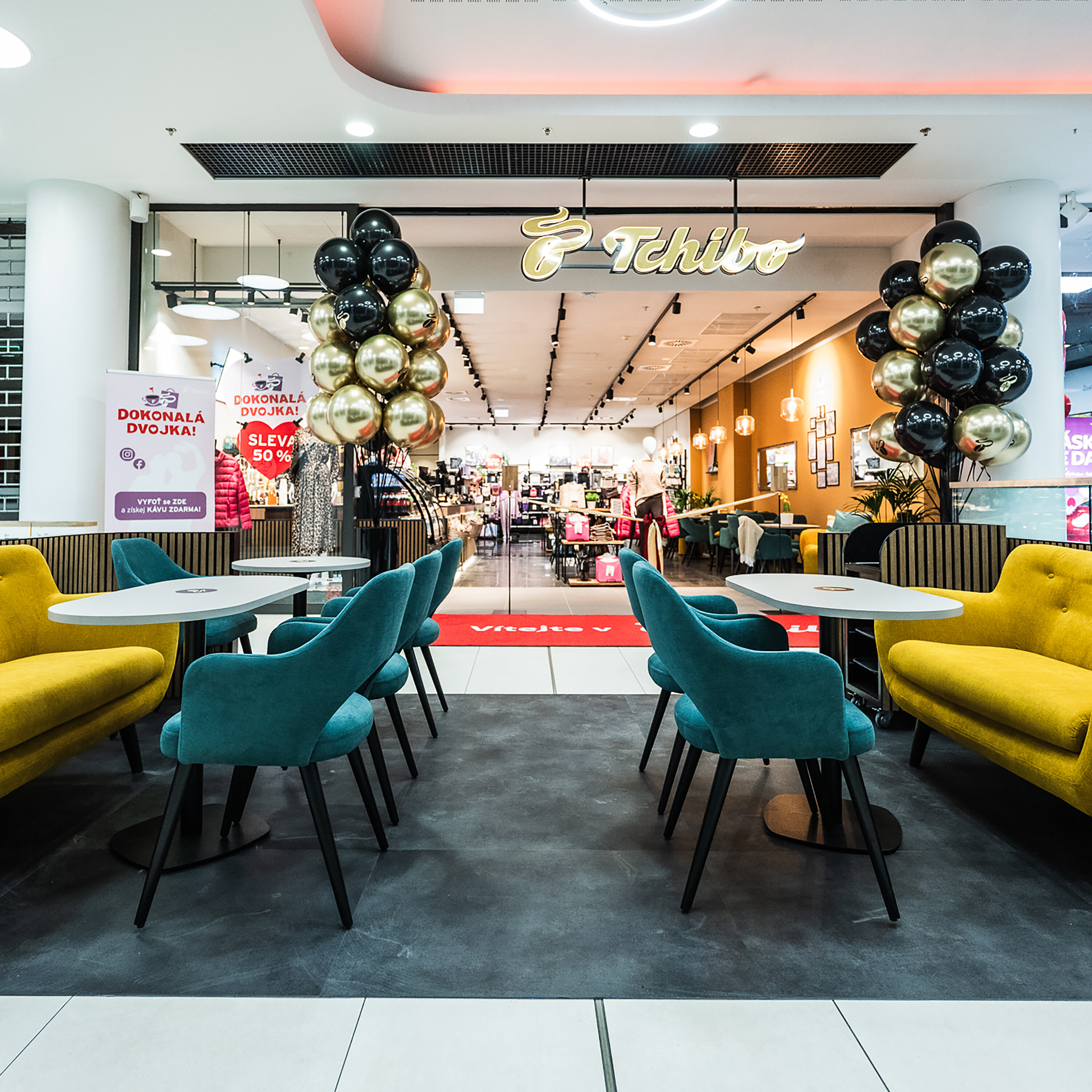 Vstup do obchodu Tchibo se žlutými pohovkami a modrými židlemi. Fasáda je zdobena balónky.