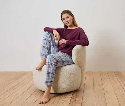 Žena sedí bosá v kostkovaných pyžamových kalhotách a vínovém topu v béžovém křesle na dřevěné podlaze.