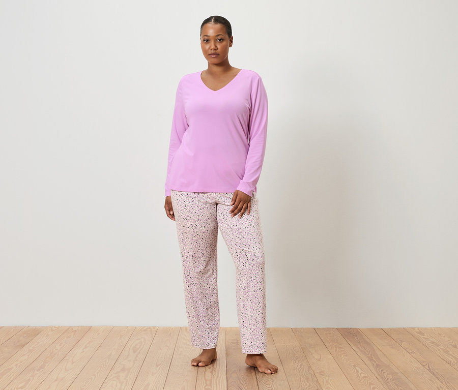 Žena stojí bosá před bílou stěnou a má na sobě fialový pyžamový top a růžové pyžamové kalhoty s květinovým vzorem.