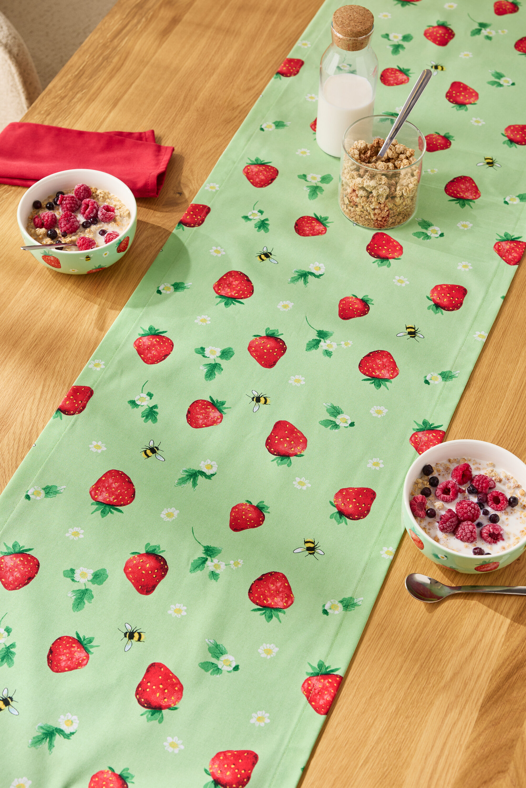 Na dřevěném stole leží běhoun se vzorem jahod, na něm misky s cereáliemi, sklenice müsli a láhev mléka.