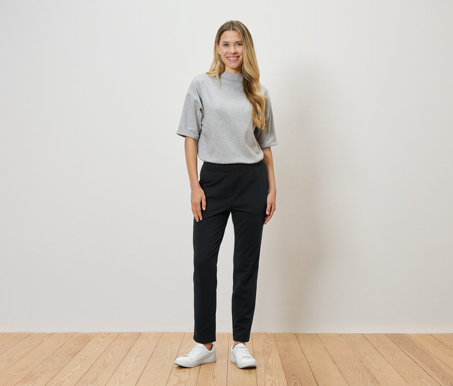 Blonďatá žena stojí před bílou zdí a má na sobě šedé tričko, černé kalhoty a bílé tenisky.