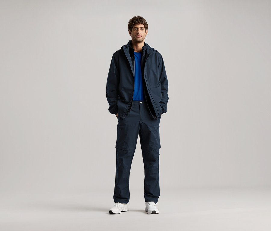 Muž stojí s rukama v kapsách tmavě modré bundy a odpovídajících kalhot, má na sobě modré tričko a bílé tenisky.