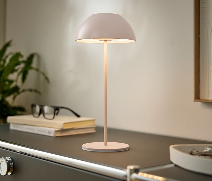 Na komodě stojí růžová lampa, knihy a brýle. V pozadí je vidět rostlina a zarámovaný obraz.