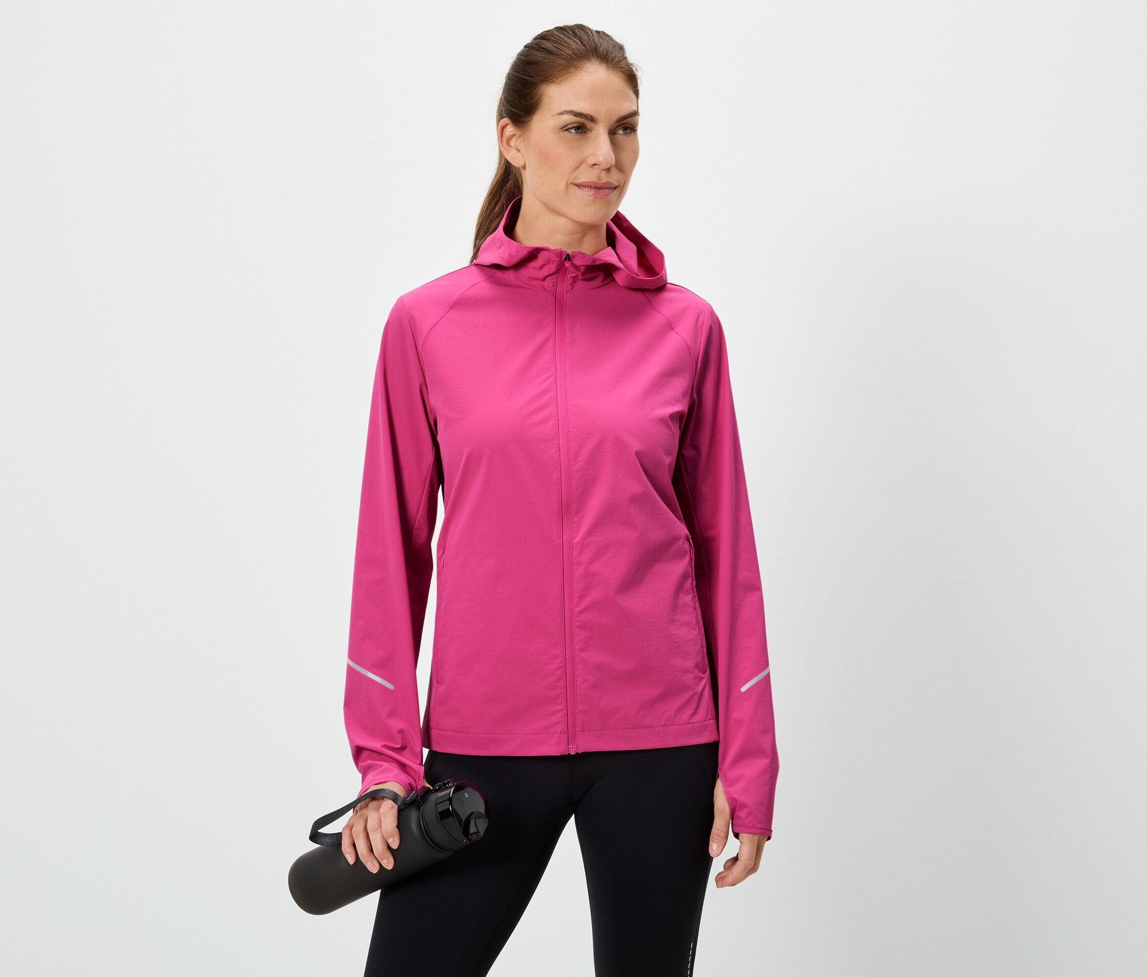 Žena drží černou láhev na vodu, má na sobě růžovou bundu a černé legíny na bílém pozadí.