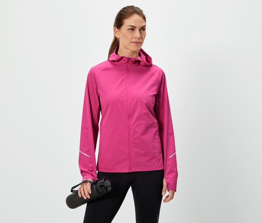 Žena drží černou láhev na vodu, má na sobě růžovou bundu a černé legíny na bílém pozadí.