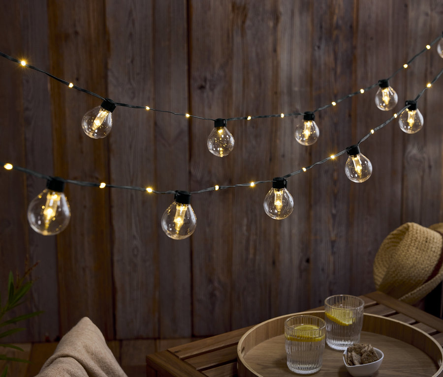 Dva řady žárovek visí před dřevěnou stěnou. Na dřevěném stole stojí dvě sklenice vody s citronem a miska ořechů.