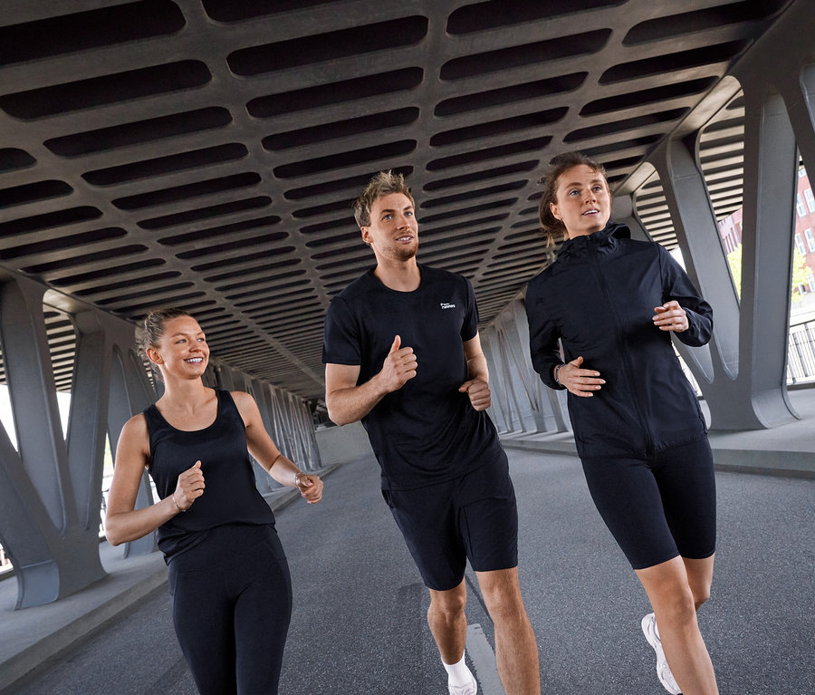 Tři lidé, dvě ženy a jeden muž, běží pod mostem. Žena vlevo má na sobě černý sportovní top a černé sportovní legíny »ActivePower«. Muž uprostřed má černé sportovní tričko a černé sportovní cyklistické šortky »ActivePower«. Žena vpravo má černou větrovku »packable« a černé sportovní cyklistické šortky »ActivePower«.