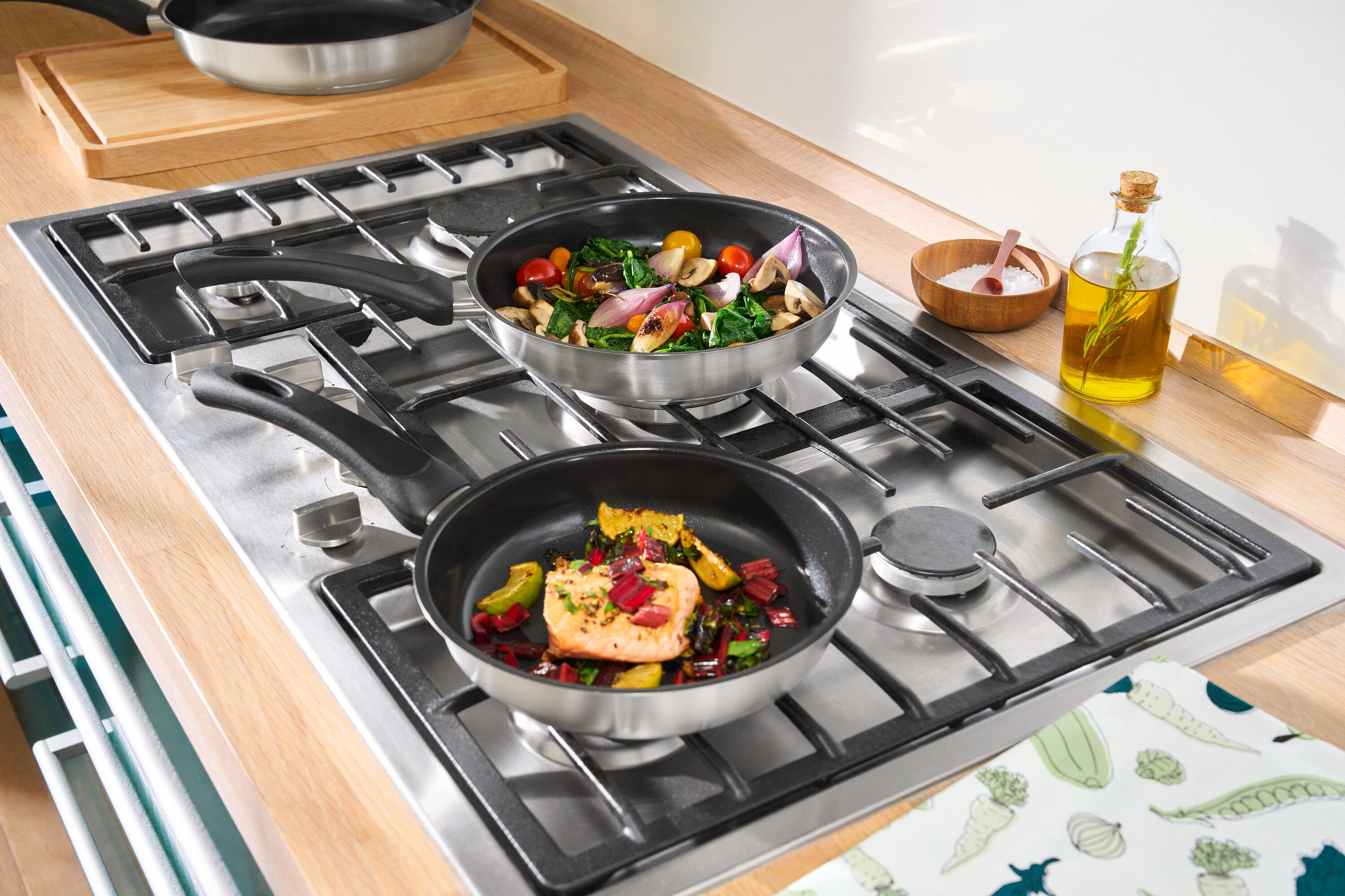 Dvě pánve z ušlechtilé oceli s keramickým povrchem na sporáku. V jedné pánvi je losos se zeleninou, v druhé zelenina.