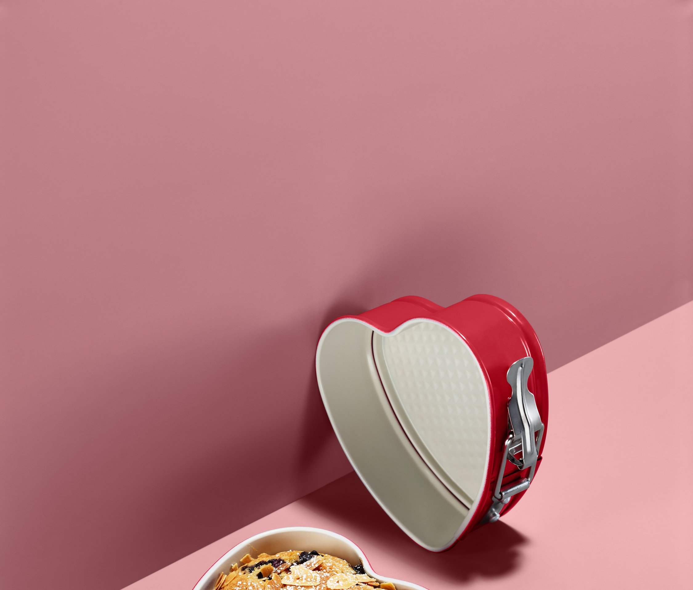 Dvě červené dortové formy ve tvaru srdce, jedna s upečeným koláčem, na růžovém pozadí.
