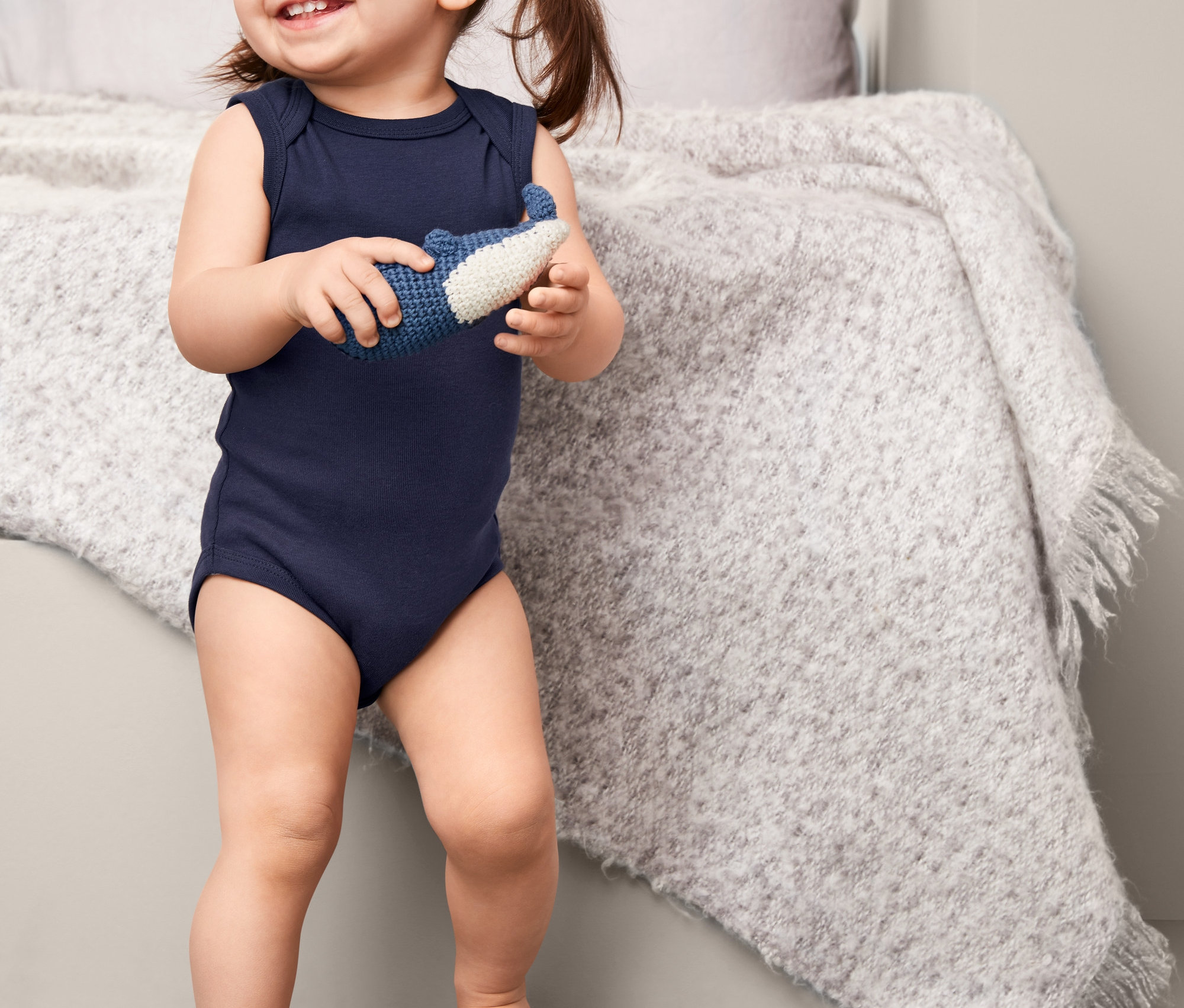 Usmívající se holčička v tmavě modrém body bez rukávů drží v rukou modro-bílou hračku.
