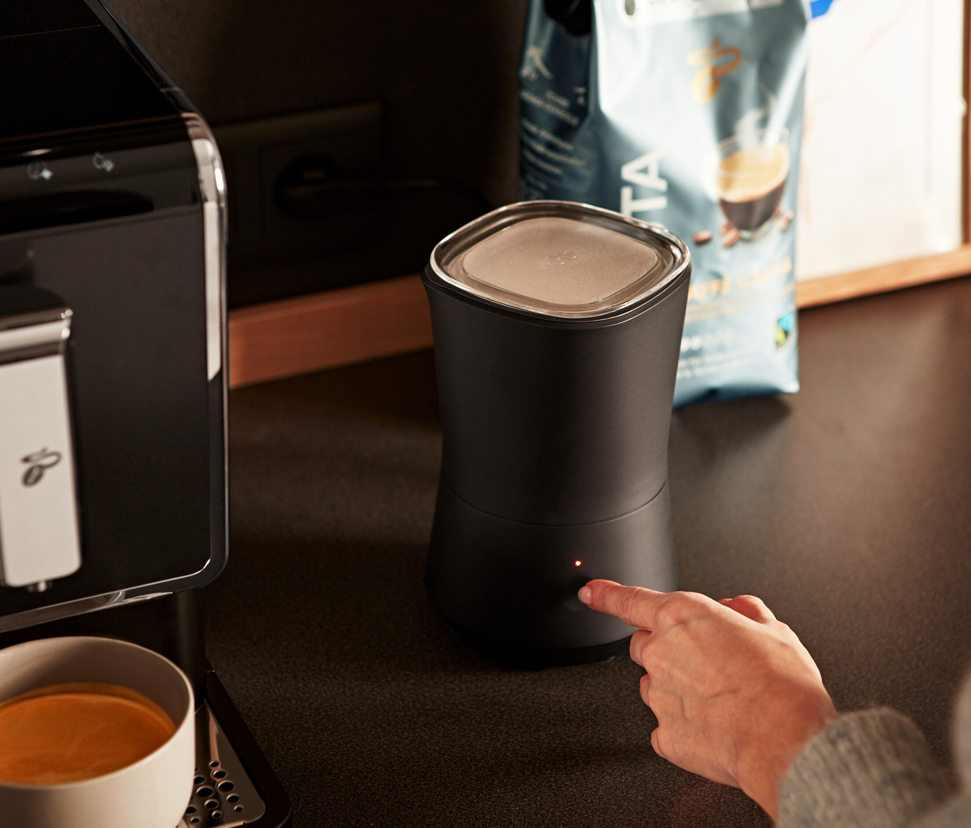 Žena zapíná antracitový elektrický napěňovač mléka. Vedle stojí kávovar a šálek s kávou.