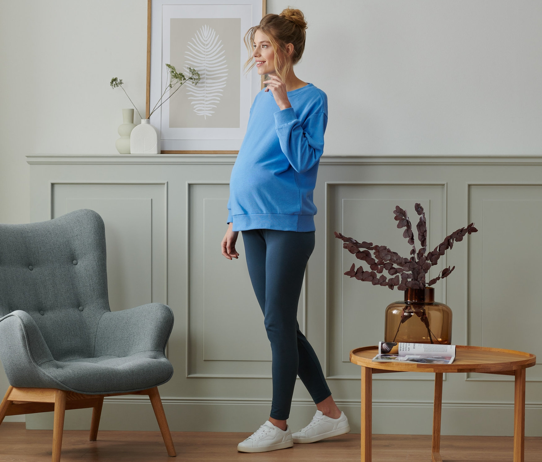 Těhotná žena stojí v místnosti a má na sobě modrý top a těhotenské legíny, 2 ks.