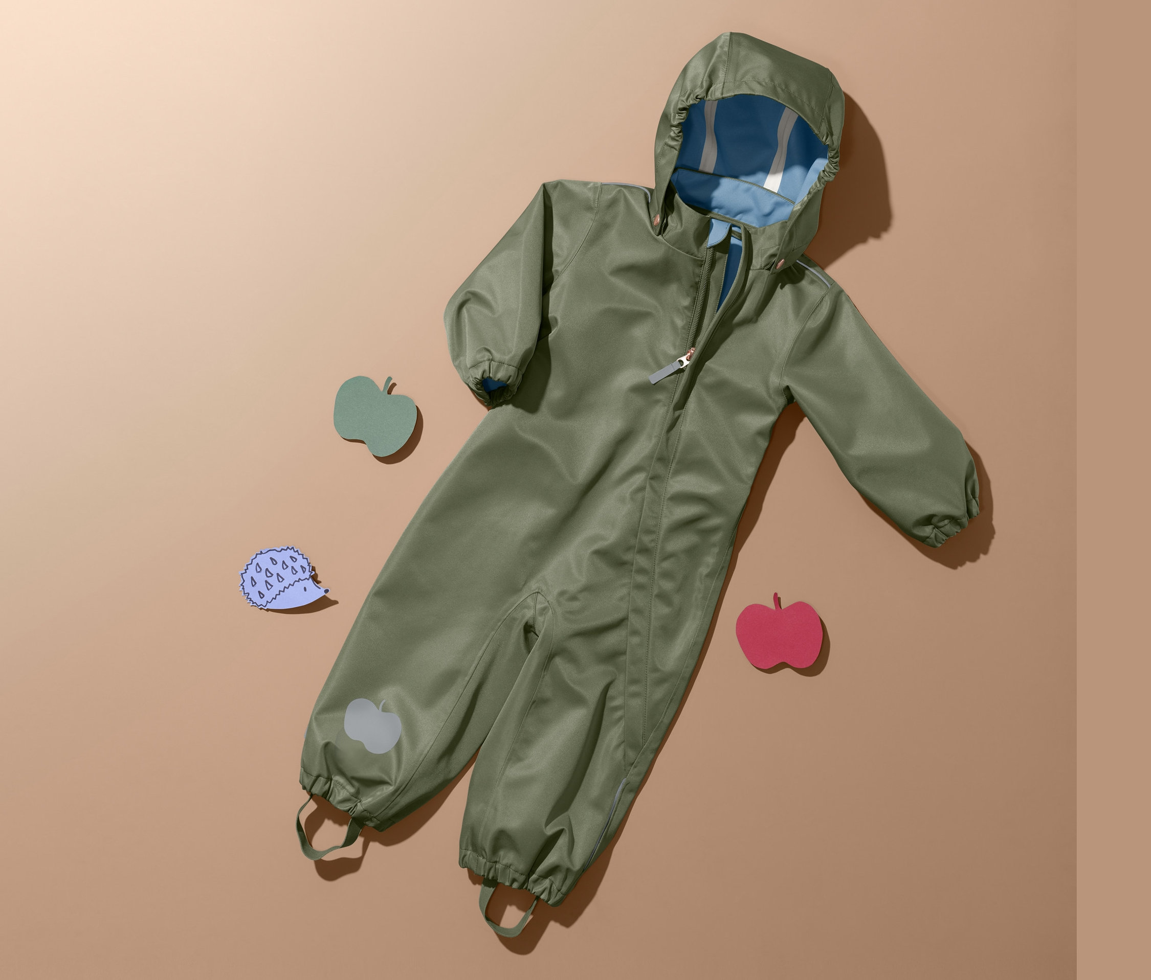 Zelená kombinéza do deště leží na béžovém pozadí vedle tří dřevěných ozdob.