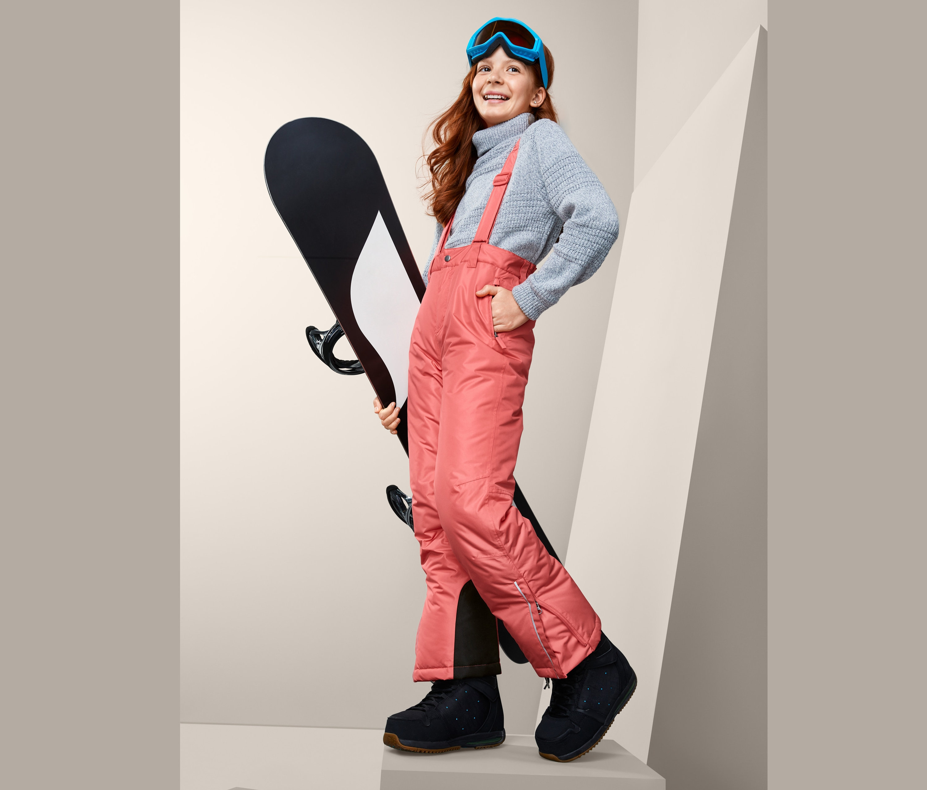 Usmívající se dívka drží snowboard a má na sobě lyžařské kalhoty, svetr a lyžařské brýle.