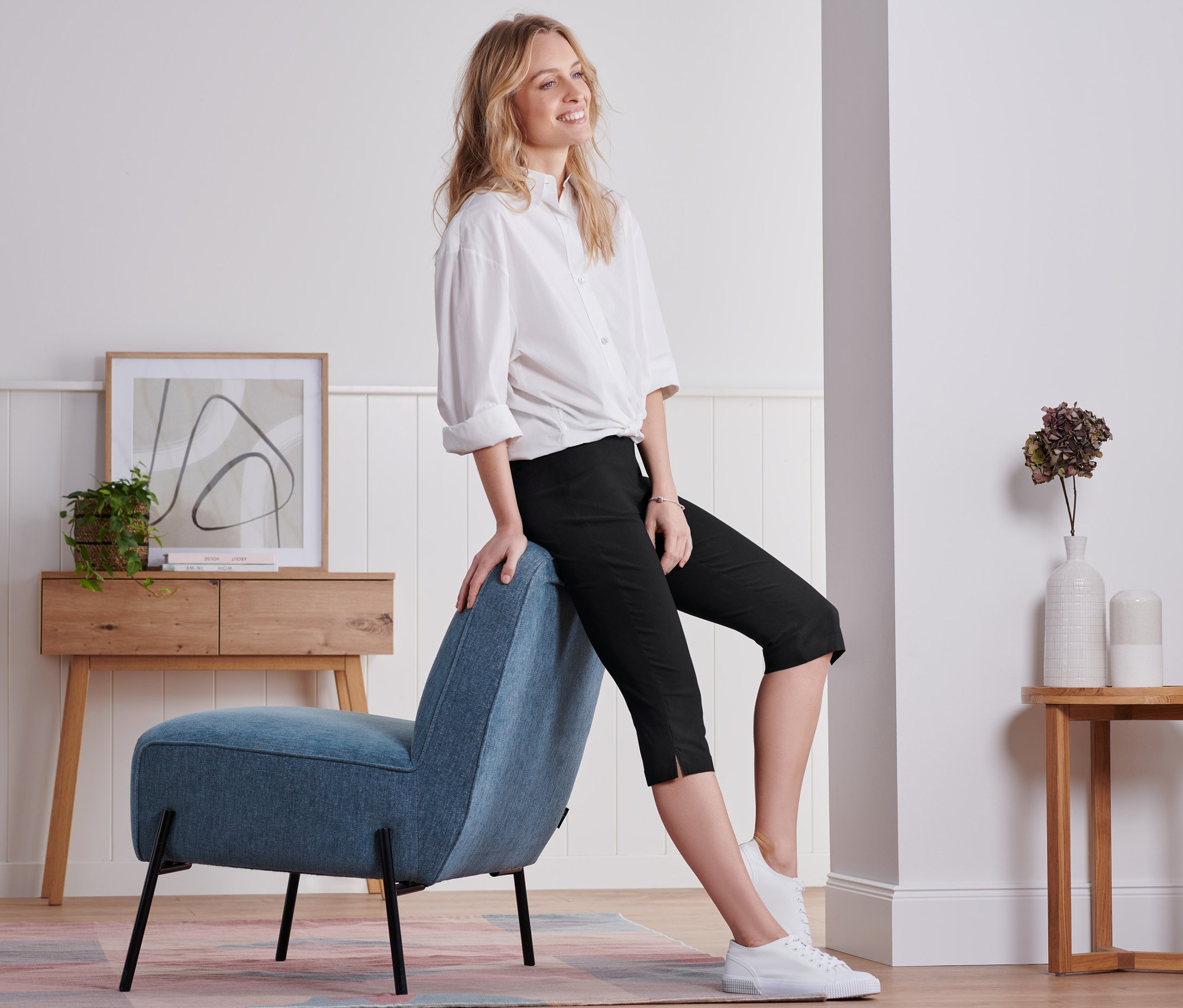 Žena sedí na modrém křesle a má na sobě černé tříčtvrteční kalhoty.