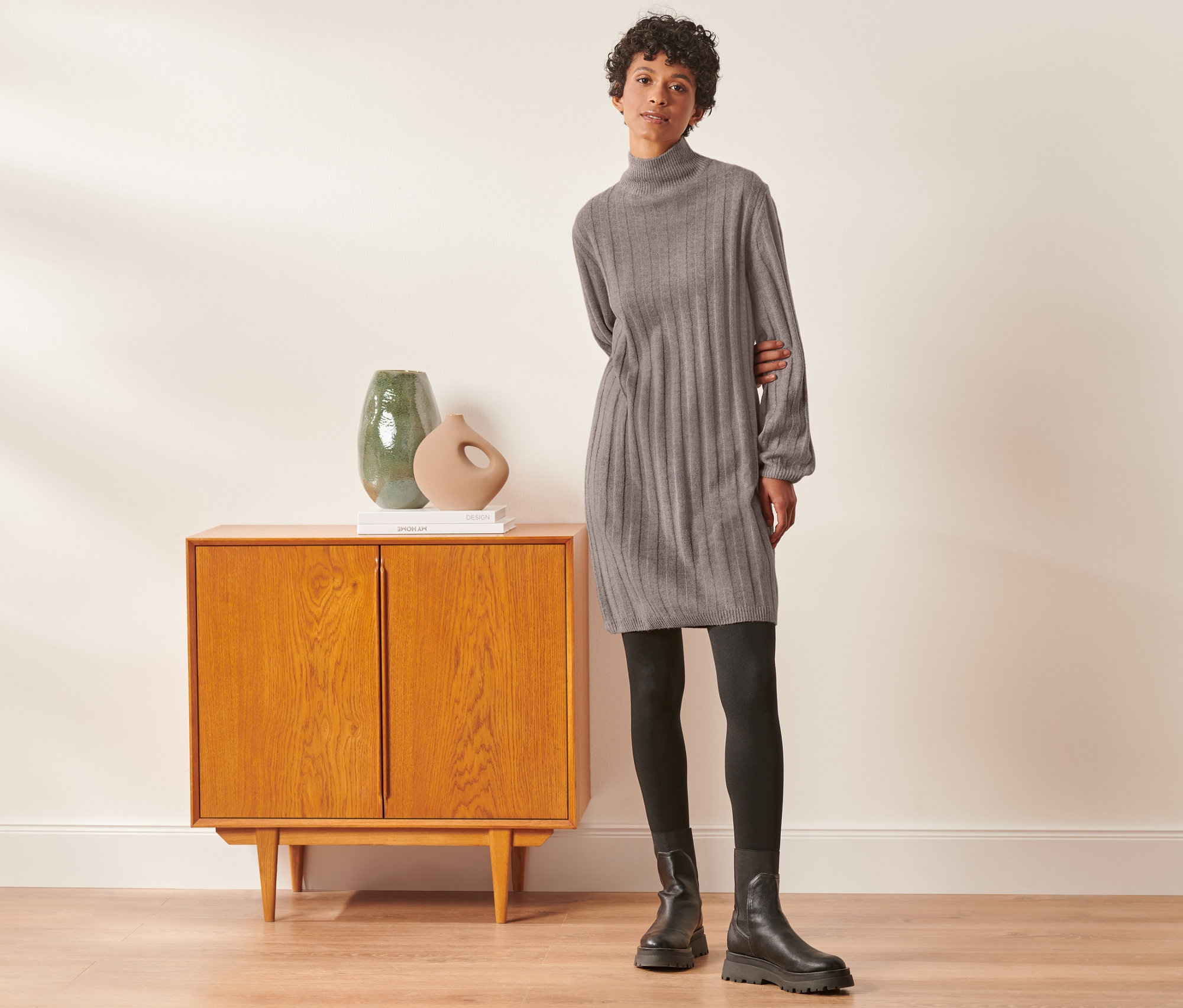 Žena pózuje vedle skříně a má na sobě šaty ze žebrované pleteniny se stojáčkem, černé legíny a boty.