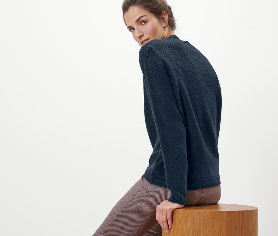 Žena sedí na dřevěné stoličce a má na sobě pletený svetr.