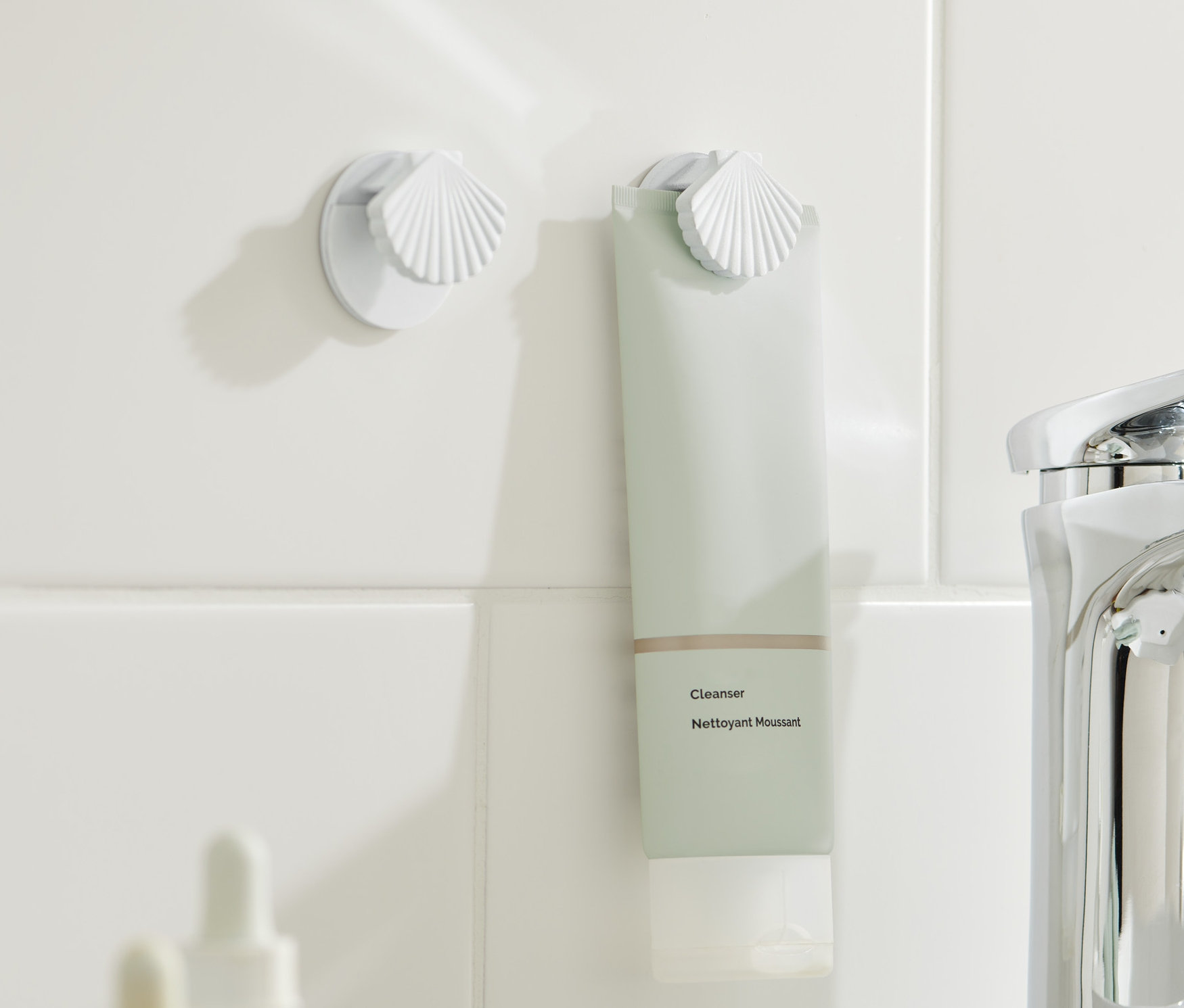 Dva držáky na tuby připevněné na bílých kachličkách, v jednom z nich je zelenkavá tuba.