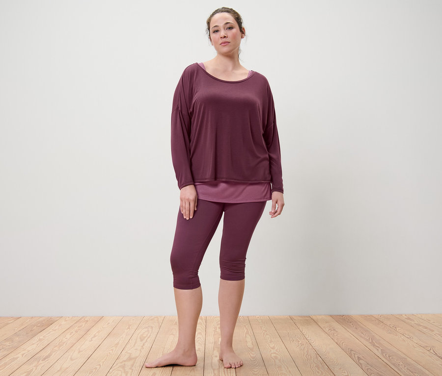 Žena stojí a má na sobě sportovní tričko 2 v 1 a 3/4 sportovní legíny ve švestkové barvě.