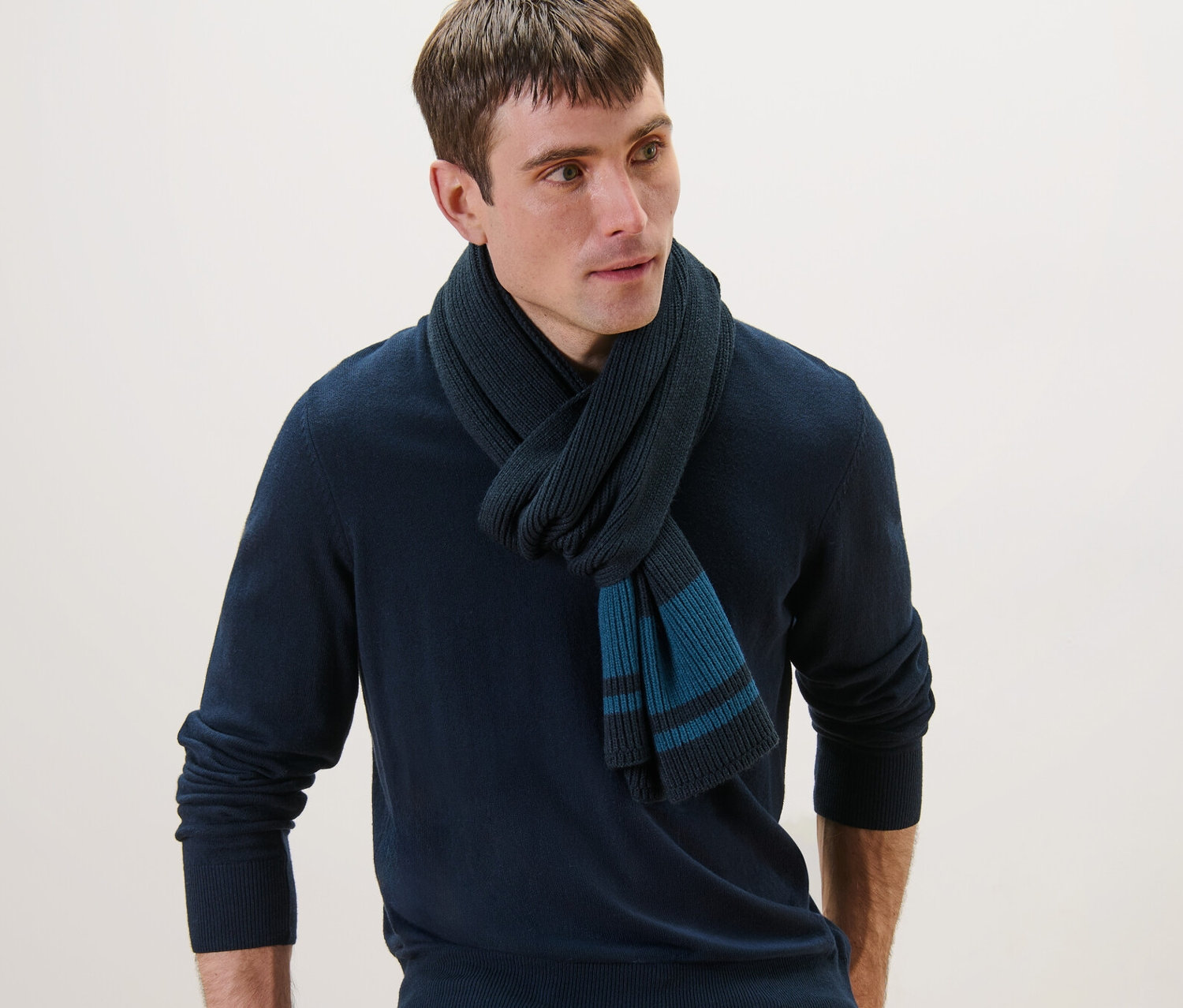 Muž s pletenou šálou, tmavě modrým svetrem a džínami sedí opřený o židli.
