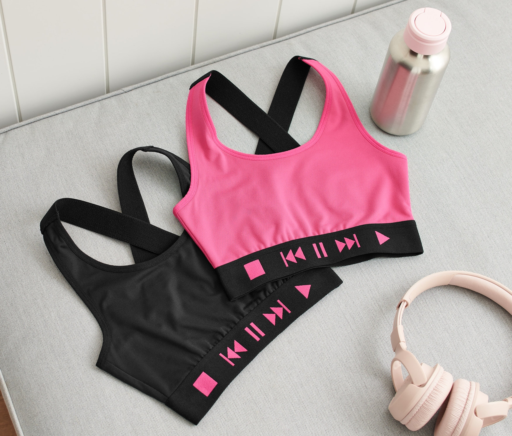 Dva dětské sportovní podprsenky, jedna růžová a jedna černá, leží na šedé pohovce vedle sluchátek, láhve s vodou a bot.