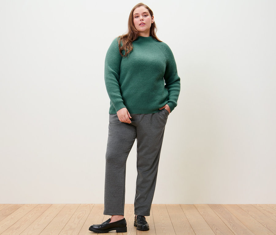 Žena pózuje v zeleném svetru z hrubého úpletu se stojáčkem a kalhotách joggpant.
