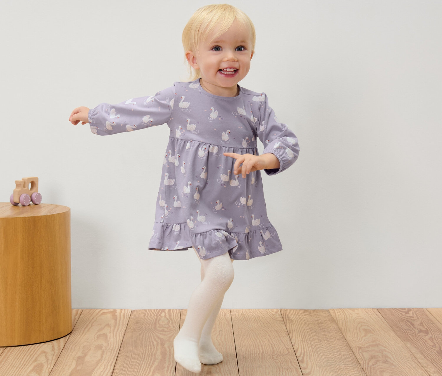 Dívka v šatech s labutěmi a punčocháčích stojí vedle dřevěného stolku s hračkou.