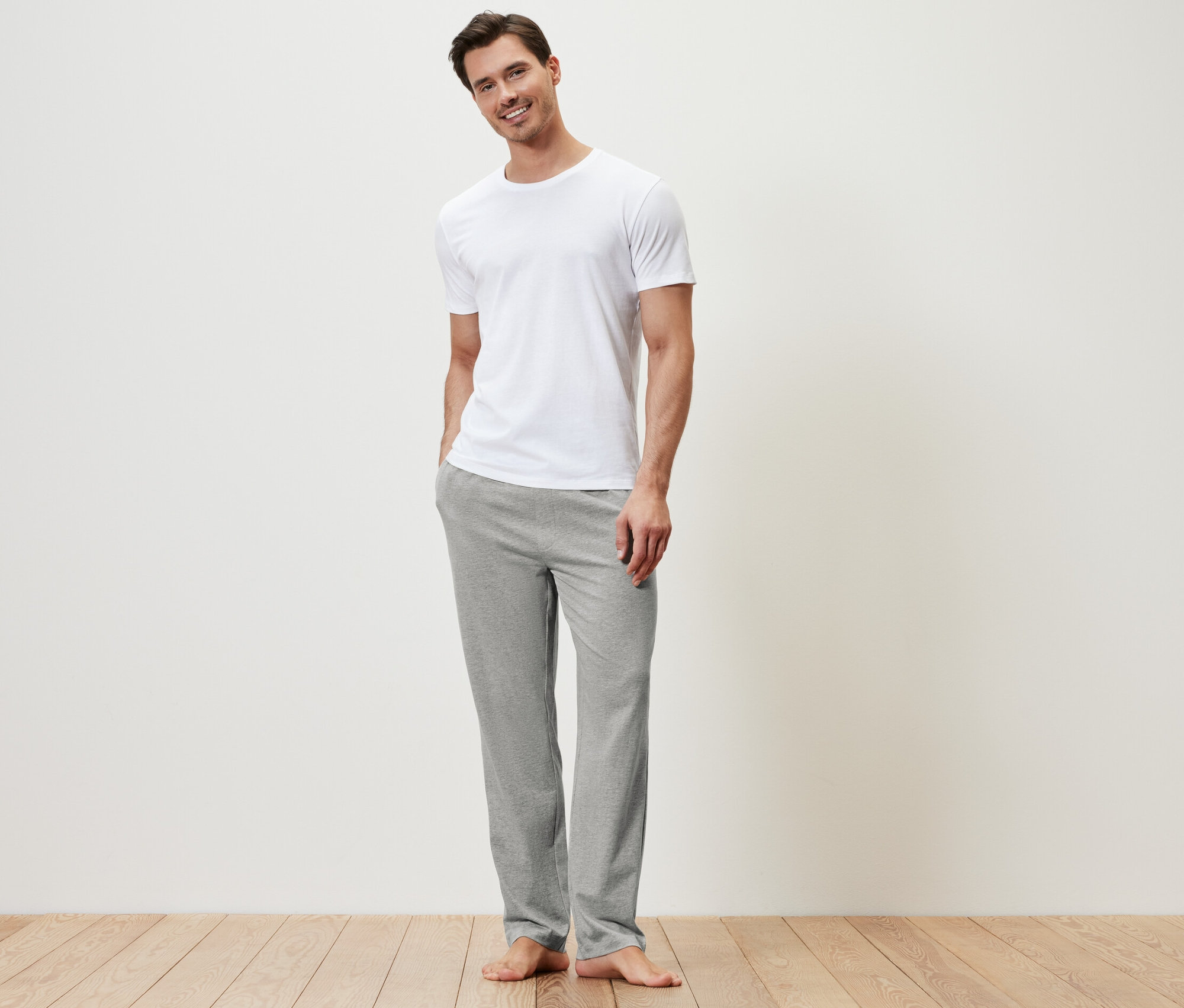 Muž stojí a má na sobě bílé tričko a šedé pohodlné kalhoty.