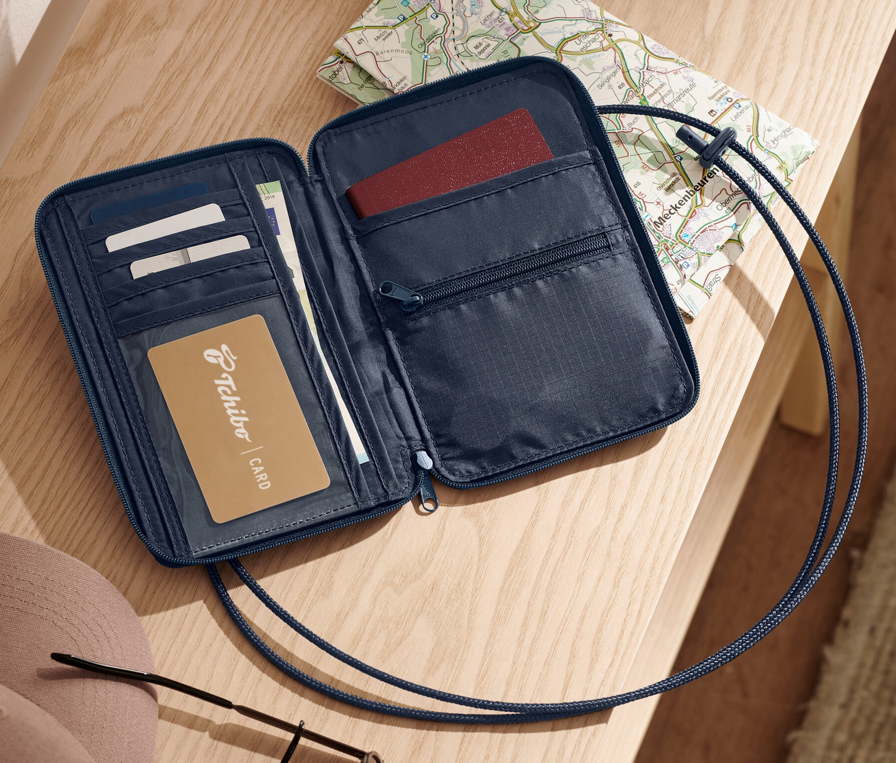 Otevřená modrá bezpečnostní náprsní taška leží na světlém dřevěném povrchu vedle mapy, klíčů, klobouku a slunečních brýlí.