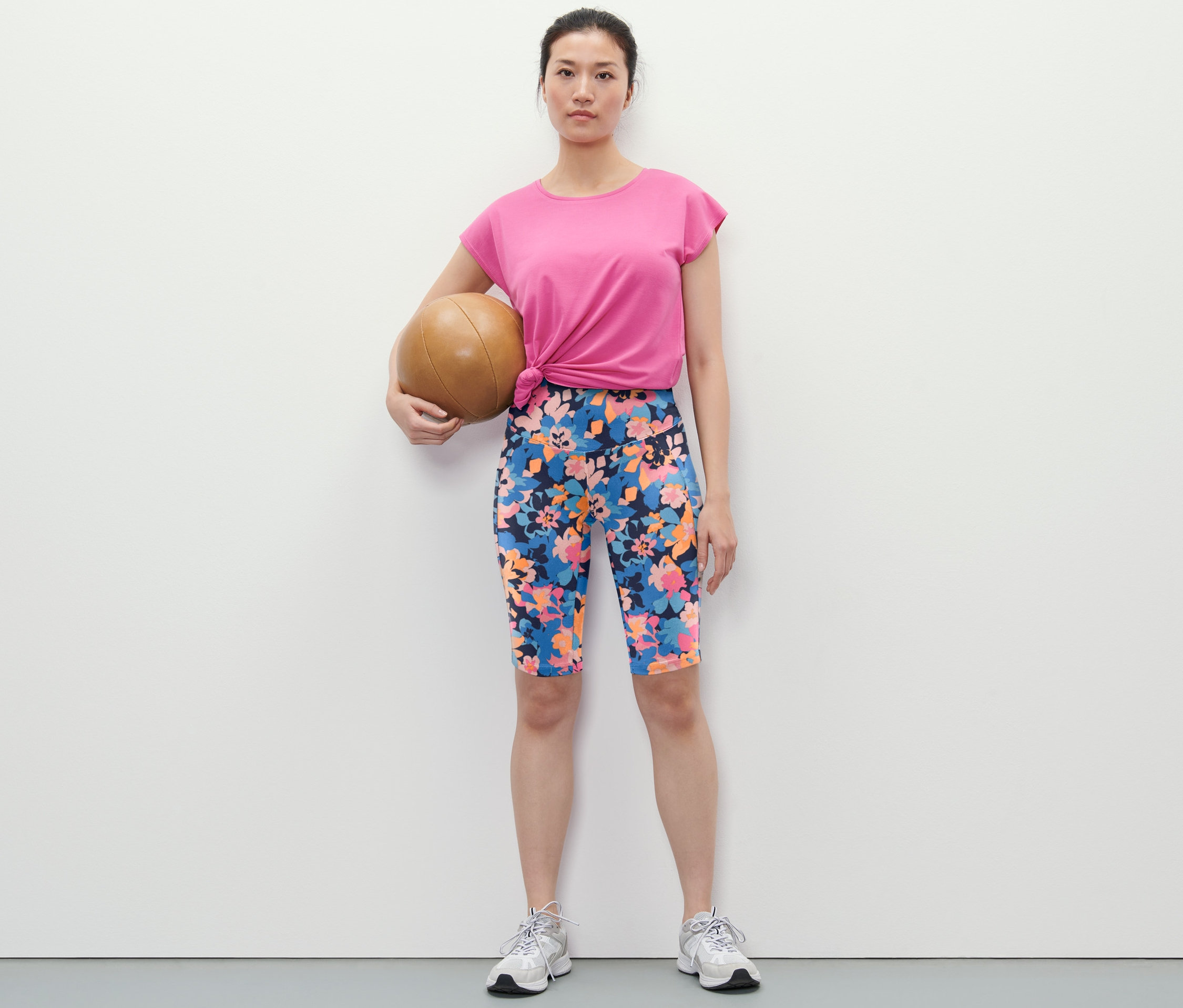 Žena drží medicinbal a má na sobě sportovní cyklistické šortky s celoplošným potiskem, tričko a tenisky.