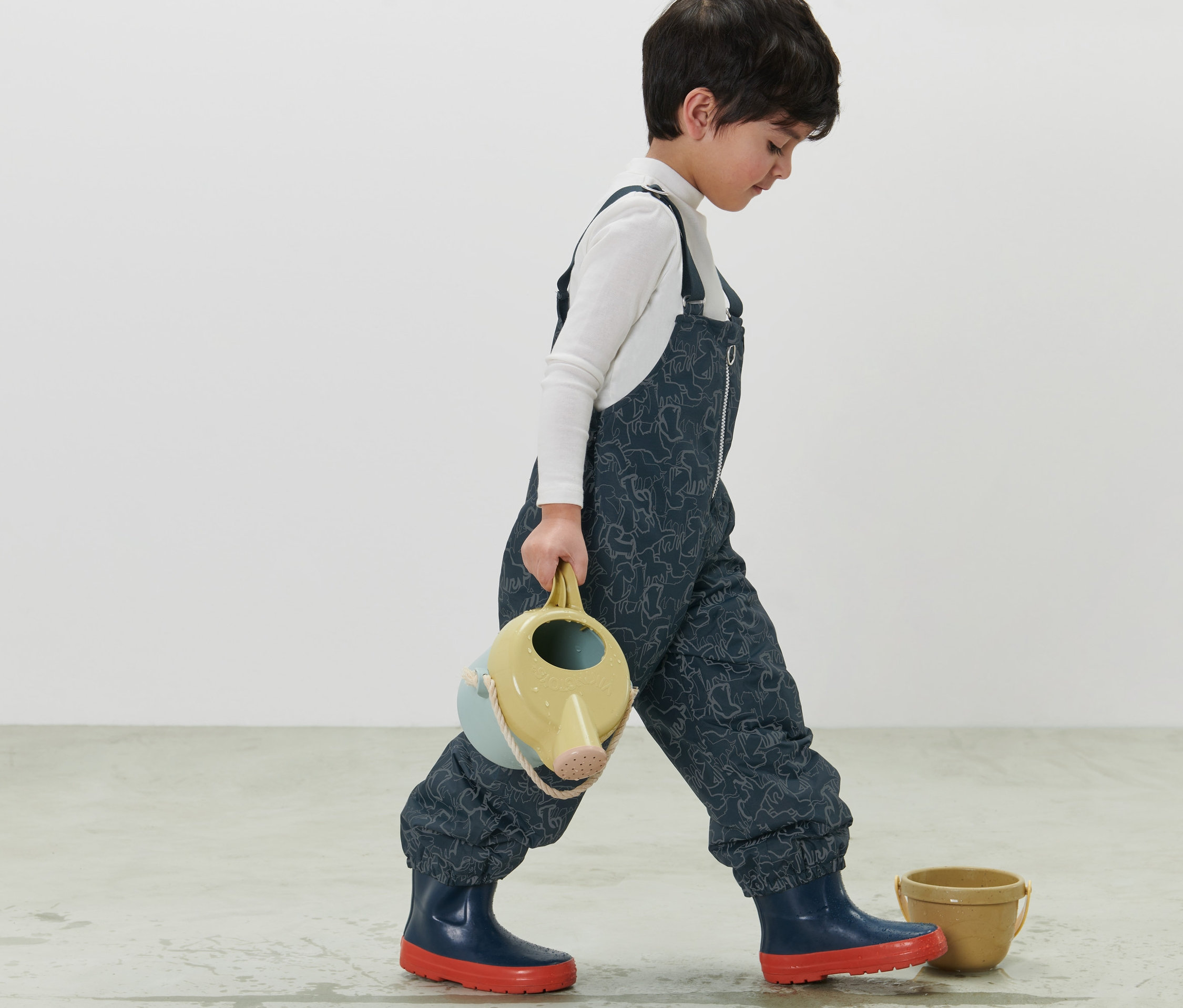 Chlapec kráčí a nese konev na zalévání. Má na sobě tmavě modré dětské termo kalhoty do deště s reflexními prvky.