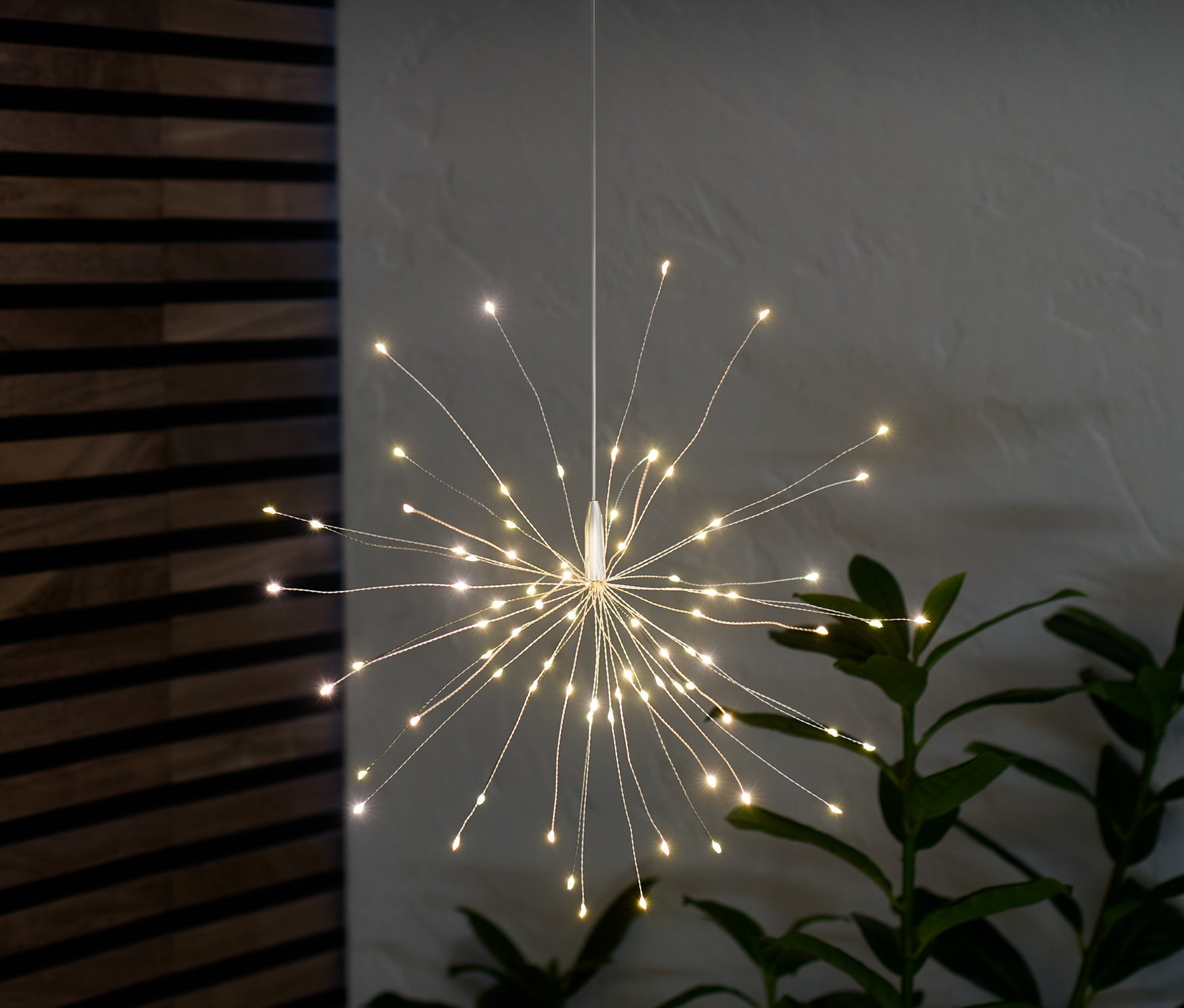 Zavěšená světelná větvička s LED diodami svítí v místnosti vedle rostliny.