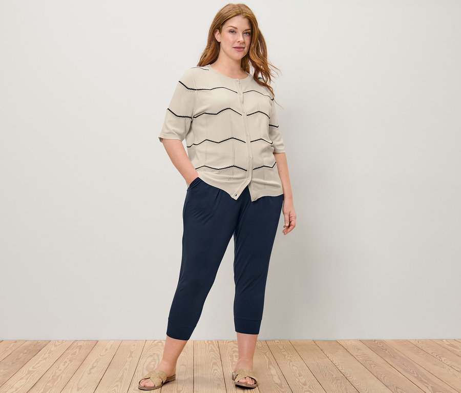 Žena pózuje v kardiganu z jemného úpletu s ažurovým vzorem a ve 3/4 žerzejových kalhotách.