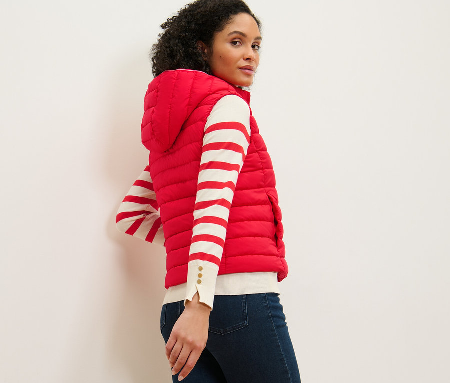 Žena se dívá přes rameno a má na sobě prošívanou vestu s kapucí, pruhovaný top a džíny.